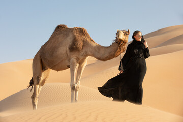 Young woman in Emirati national dress (abaya) with a camel in Empty Quarters desert dunes. Abu...