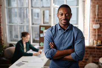 Young successful African-American businessman with arms crossed by chest standing against female colleague working in front of laptop