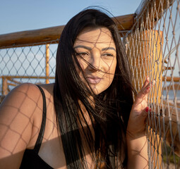 Brunette model with calm expression posing in the shade of a hammock on a beach access walkway with sun on her face and a blue sky. Mention of casual, vacation and photographic poses.