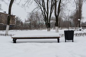 A bench, in the park, next to a trash can, sprinkled with snow. Winter park, strewn with snow, with lanterns and trees.