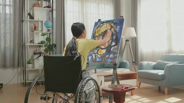 Hind View Of An Asian Artist Girl In Wheelchair Holding Paintbrush And Painting A Girl'S Face On The Canvas
