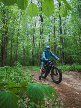 An adult man on an electric bike with thick wheels. The concept of outdoor sports in any weather. Fat bike in the forest in the rain.
