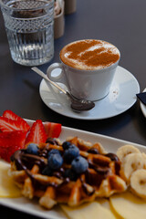 Traditional Italian breakfast. Fresh classic waffles with fruits, sugar powder, chocolate and cappuccino coffee with cinnamon in Milan, Lombardy, Italy. European food, drinks  pastry at a local bar.