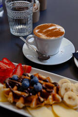 Traditional Italian breakfast. Fresh classic waffles with fruits, sugar powder, chocolate and cappuccino coffee with cinnamon in Milan, Lombardy, Italy. European food, drinks  pastry at a local bar.