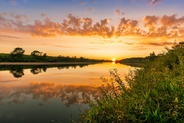 Naklejka na ściany i meble Scenic view at beautiful summer river sunset with reflection on water with green bushes, calm water ,deep colorful cloudy sky and glow on horizon on a background, spring evening landscape