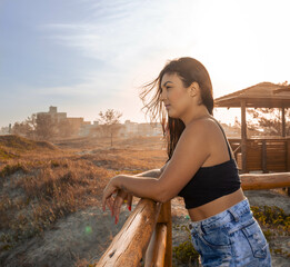 Girl wearing jeans and black crop top contemplating the sea on an access walkway near dunes with the sun in the background and a blue sky of a vibrant day. Mention of holidays, rest and relaxation