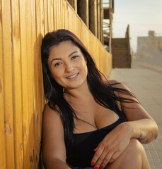 Brunette girl sitting wearing jeans and black crop top contemplating the sun on a walkway to the sea resting propped up and a blue sky of a vibrant day. Mention of holidays, rest and relaxation