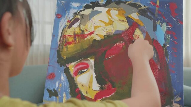 Close Up Of An Asian Artist Girl Sitting On Round Wooden Chair Without Backrest Holding Paintbrush Mixed Colour And Painting A Girl'S Face On The Canvas
