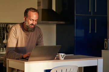 Portrait of a smiley middle-aged bearded man using his laptop early in the morning at home