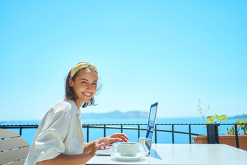 Beautiful young mixed race woman smiling to camera enjoying using laptop online internet at outdoor resort cafe with sea view