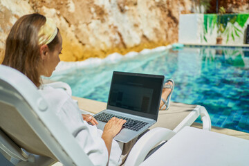 Faceless female using laptop computer online internet while lying on sun loungers by swimming pool...