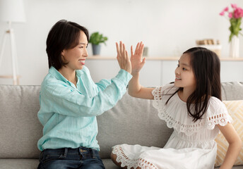 Cute little Asian girl giving high five to her grandmother, sitting on couch at home. Loving multi...