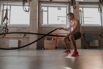 Side view of handsome physically fit male working out with heavy ropes in cross fit gym