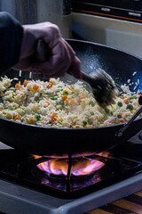 Woman's hand is a blur as she stirs the rice and veggies in the cast iron wok on the gas stove. with a large wooden spoon.