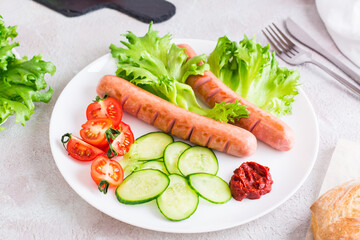 Ready to eat fried sausages, chopped tomatoes and cucumber vegetables on a plate and a bun on the table. Hearty homemade snack
