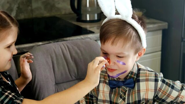 A girl in bunny ears paints a boy's nose in the kitchen at home. The boy laughs. Fun, Easter. Close-up