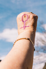 Fist of an older woman with purple painted nails, with the sky in the background, with the female symbol painted. Concept of women's day, empowerment, equality, inequality, activism and protest.