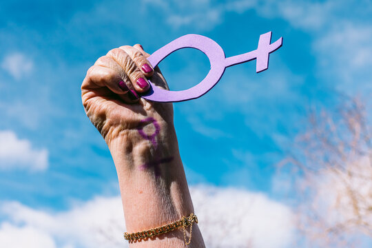 Hand of an older woman with purple nails, with the sky in the background, holding the female symbol in purple. Concept of women's day, empowerment, equality, inequality, activism and protest.