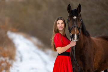 Fototapeta premium Portrait of a beautiful girl in a red dress standing with a horse on the background of a winter road