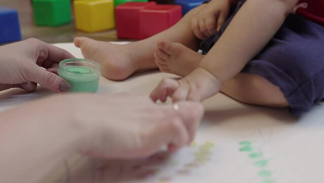 mother teaches toddler baby painting with finger paints on white paper. child sitting on floor taking paint bottle in hands mom showing how put finger in paint and color paper. kids home education 