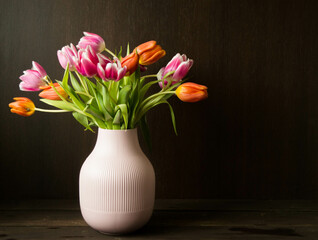 pink and orange colored tulips in vase in front of a dark brown wooden wall