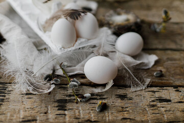 Stylish Easter rustic still life. Easter natural eggs in tray, feathers, willow branches, nest, linen cloth on aged wooden table. Happy Easter! Simple rural aesthetics