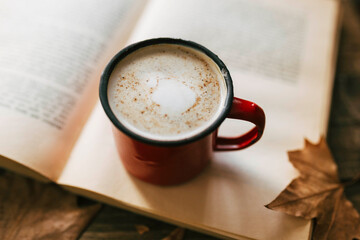 Enamel mug of milk with cinnamon. Reading a book in autumn. Wooden table with autumnal leaves. Morning routine.. Focus on cocoa and cinnamon.