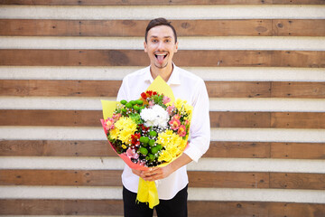 Fototapeta premium Summer, flowers, emotions and people concept - Satisfied Caucasian guy in a white shirt and black jeans showing tongue and holding a big bouquet on brown ribbed background with copy space