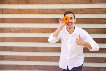 Funny Caucasian guy in a white shirt showing thumbs up and tongue holding an orange flower instead of an eye on a brown ribbed background with copy space