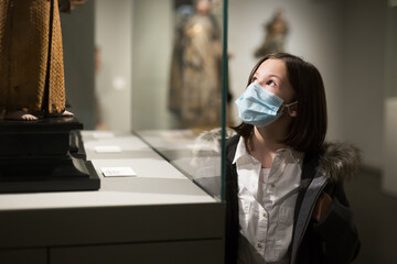 Cute interested preteen girl wearing protective face mask exploring artworks in modern museum of...