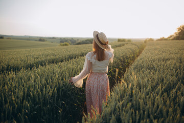 Young girl in a beautiful long dress and hat walks in a field with wheat in his hand © sergiokat