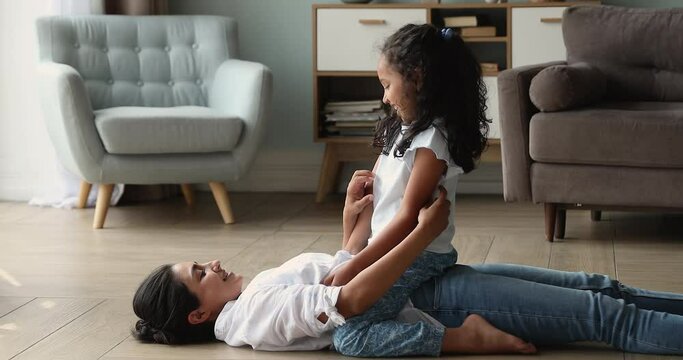 Indian woman lying on warm floor tickles her cute little cheery daughter. 6s girl have fun enjoy playtime with loving mum or nanny, spend carefree playful time together at home. Leisure, bond concept