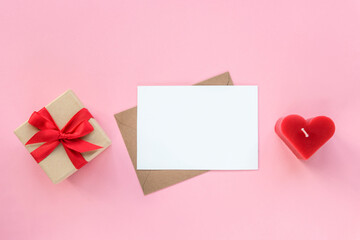 Gift box, envelope with blank card and red candle in a shape of heart on pink background. Valentines day concept. Top view, flat lay, mockup