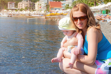 Fototapeta na wymiar A smiling mother with a small baby on the seashore. Beach leisure with children. Summer time.