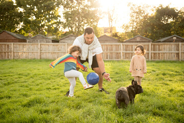 A father holds a football as her daughter kicks it off his hands while her toddler friend and dog...