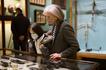 Interested preteen girl and elderly woman wearing protective face masks viewing antique handguns...