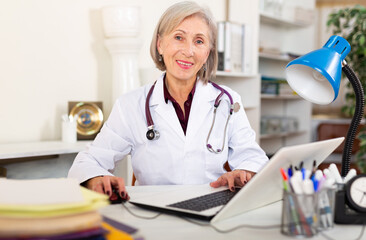 Portrait of friendly smiling elderly female doctor giving consultation in clinic office, sitting at table with laptop