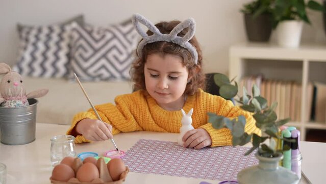 Happy little girl with bunny ears painting the clay rabbit preparing for Happy Easter day. Preparing handmade Easter decoration at home.