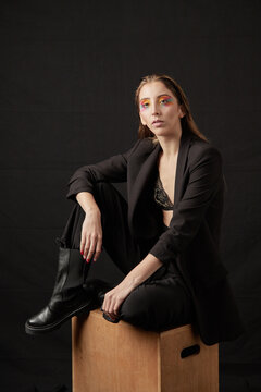 Portrait of a young blonde model, sitting on a brown wooden box, wearing a black dress. In studio on black background.