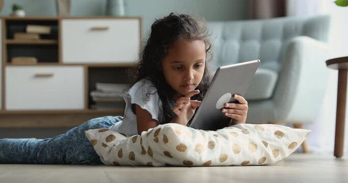 Adorable preschool Indian girl lying in living room on warm floor using digital tablet, play video games, watch favourite vlogger, spend free time alone at home. Gen Z use modern wireless tech concept