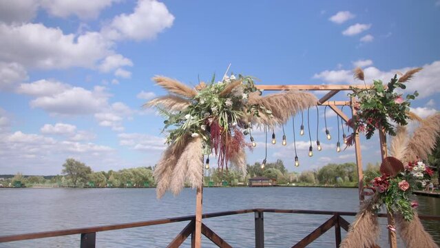 A beautiful wedding arch decorated with flowers stands in front of the pond on a warm summer day. a pier with rustic decor stands near the blue lake