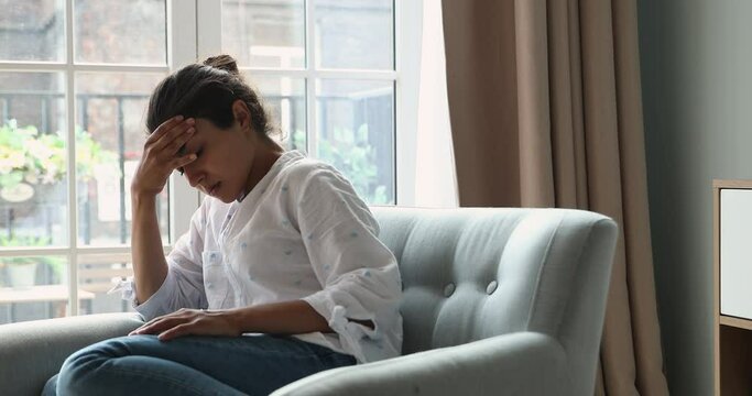 Worried Indian woman sit on armchair looks concerned, feels depressed due life troubles break up, divorce, thinks about abortion suffering alone at home. Psychological problem, regret, mistake concept