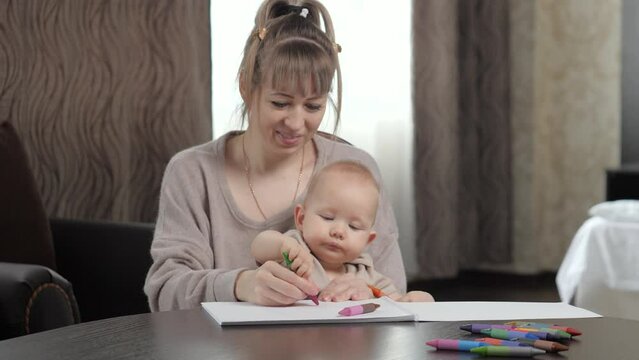 Mom, baby play together and draw at home in room. Young mother teaches cute baby son to draw with colored markers on paper sheet. Family adheres to early education sitting at table in bedroom at home.