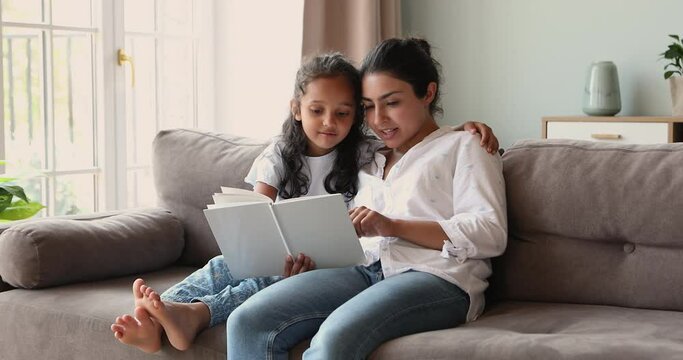 Cheerful Indian woman read fairytale book to her cute little daughter sit together on couch at home. 6s girl enjoy fascinating story, listen mommy storytelling. Kid development and upbringing concept