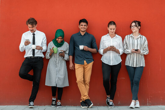 Multiethnic group of casual businesspeople using smartphone during a coffee break from work in front of the red wall outside.