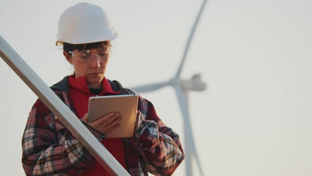 Young woman engineer in helmet works with a tablet computer on wind turbine power generator station. Windmills on fied. Slow motion