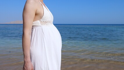 Fototapeta na wymiar Beauty of pregnancy period, woman with big baby bump in white dress walking on the sunny ocean coast 