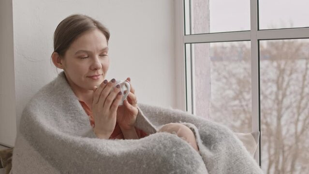 Medium slowmo shot of young dreamy woman in cozy gray plaid sitting with cup of hot tea on windowsill looking at window