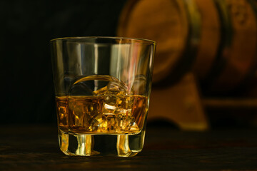 A glass of whiskey with ice cubes against the background of an oak barrel. Close-up. Dark background.