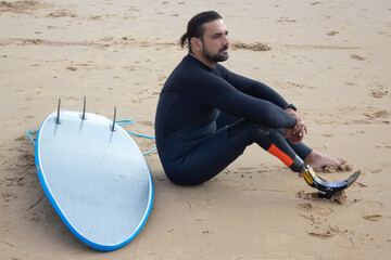 Dreamy mid adult man resting after training on beach. Man with beard and dark hair in surfing suit sitting on sand, resting. Sport, leisure, disability , active lifestyle concept © KAMPUS